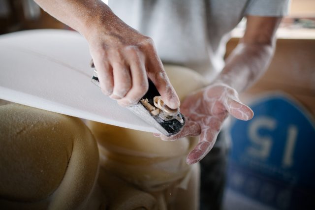 Close up photo of artist shaping a surfboard inside workshop